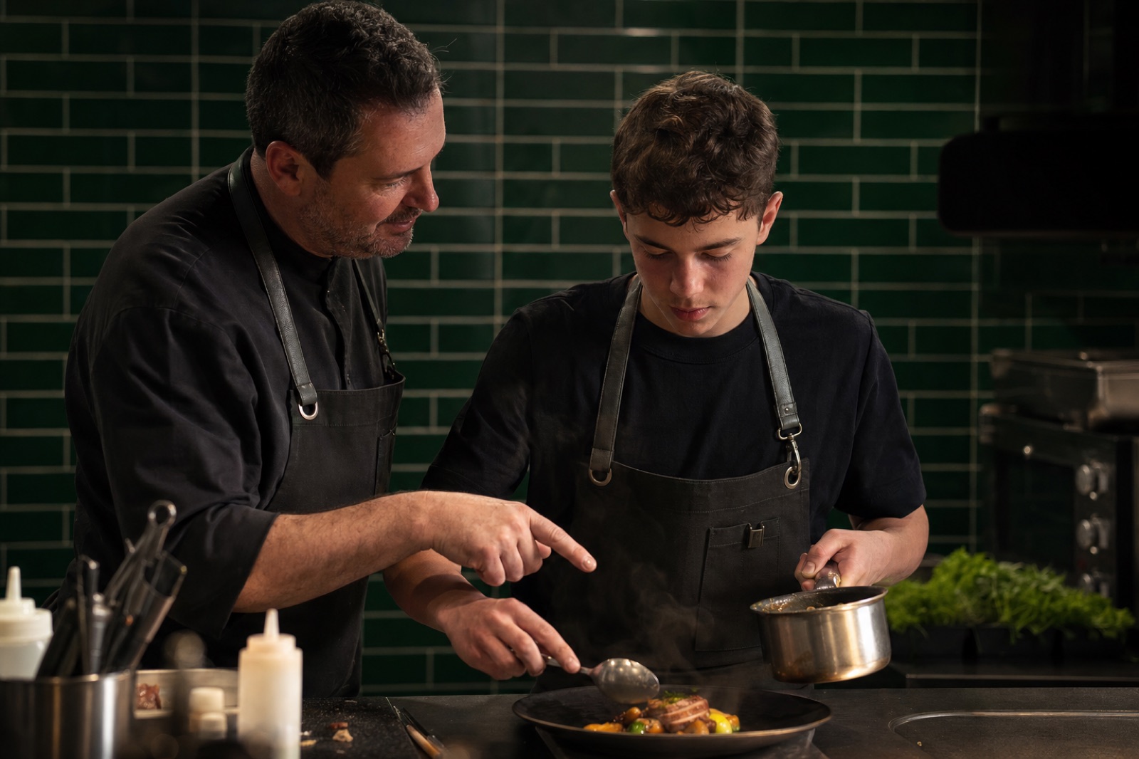 A chef mentoring a young culinary student at the stove — craft passed forward